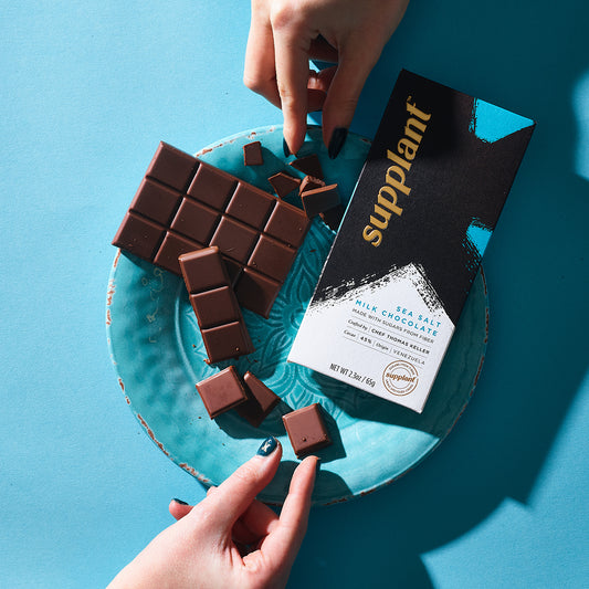 Front of pack of sea salt milk chocolate bar on plate with hands holding chocolate on blue background
